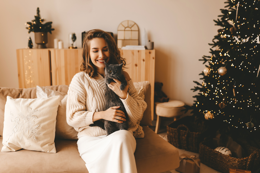 woman in knitted cozy clothes hugs her cat sitting on the sofa near the decorated Christmas tree