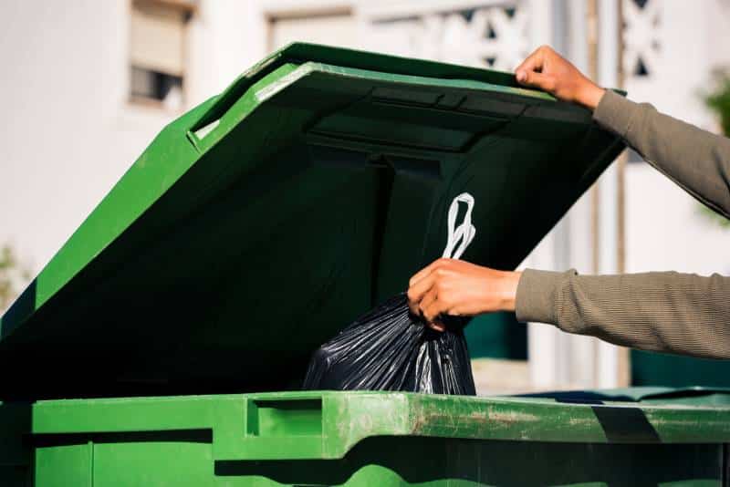 Man throwing out black eco-friendly recyclable trash bag in to big plastic green garbage container
